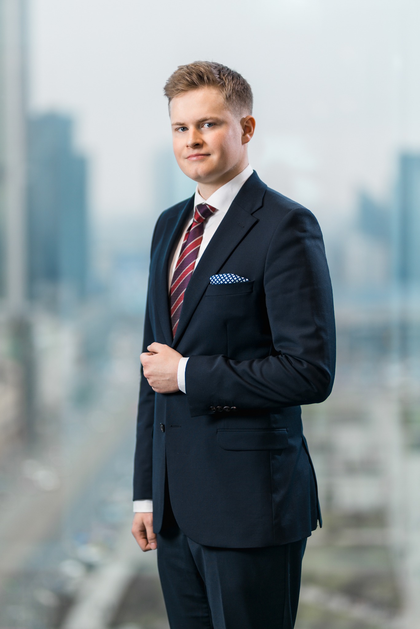 Portret młodego mężczyzny w granatowym garniturze, czerwono-bordowym krawacie w paski i poszetce w niebieskie kropki, pozującego na tle okna z widokiem na miasto.