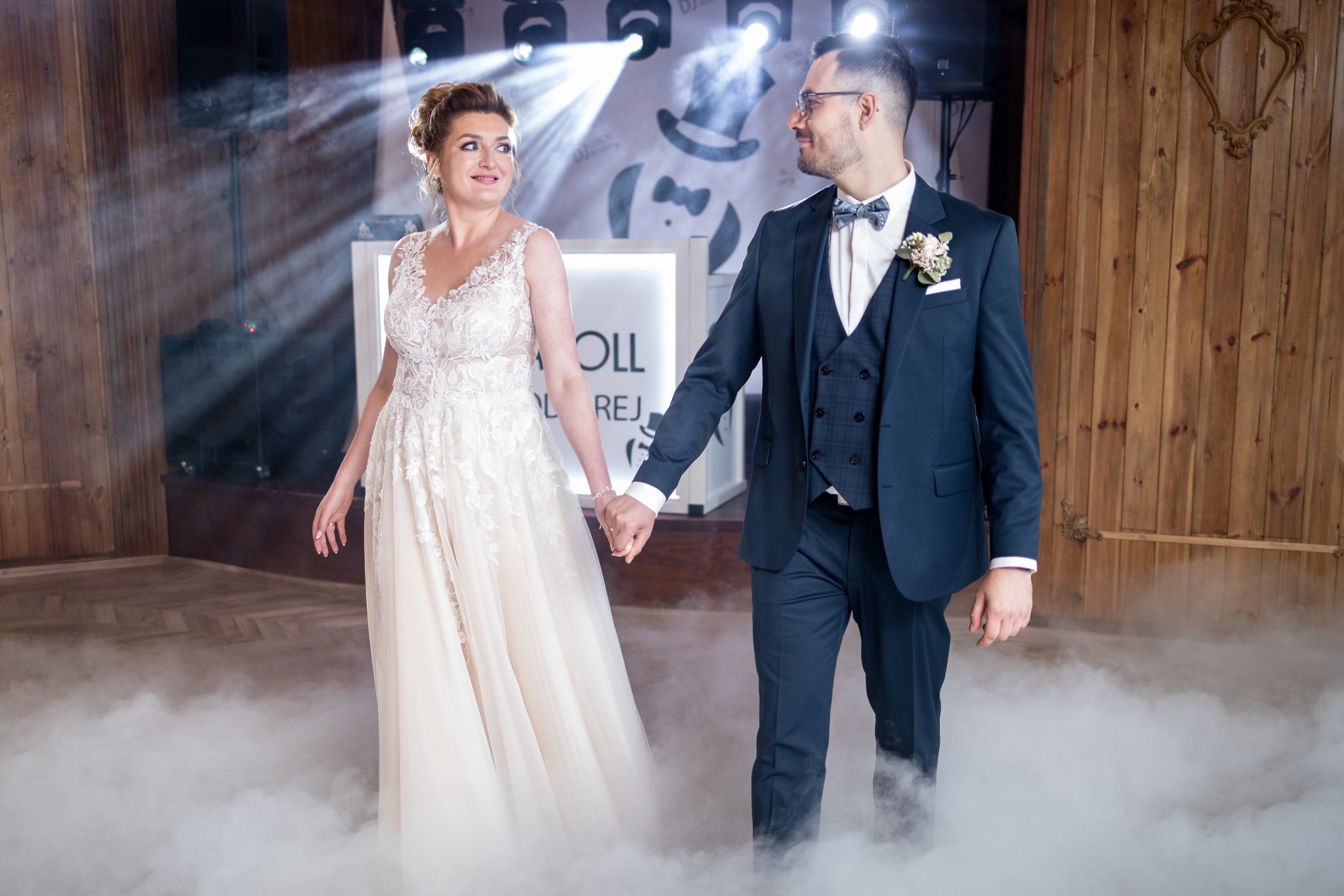 Para młoda, on w granatowym garniturze z butonierką, ona w koronkowej sukni ślubnej, trzymają się za ręce na parkiecie spowitym mgłą, w tle drewniana ściana i oświetlenie sceniczne.