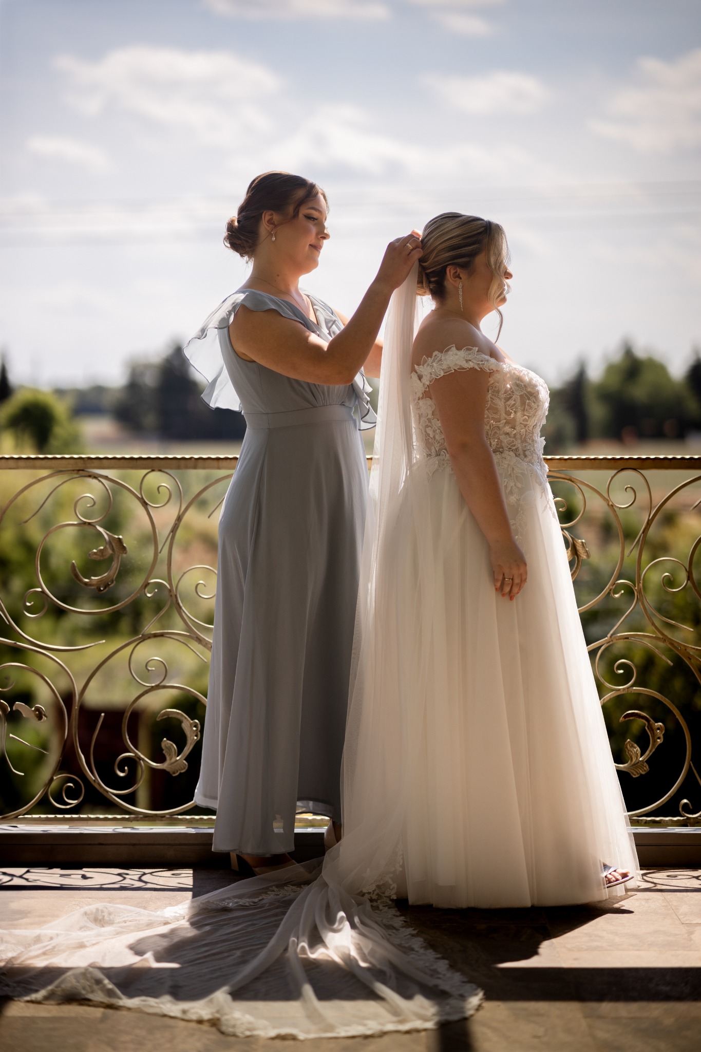 Panna młoda w koronkowej sukni ślubnej i welonie, poprawiana przez druhnę w szarej sukience na balkonie z ozdobną balustradą, z widokiem na zielone drzewa.