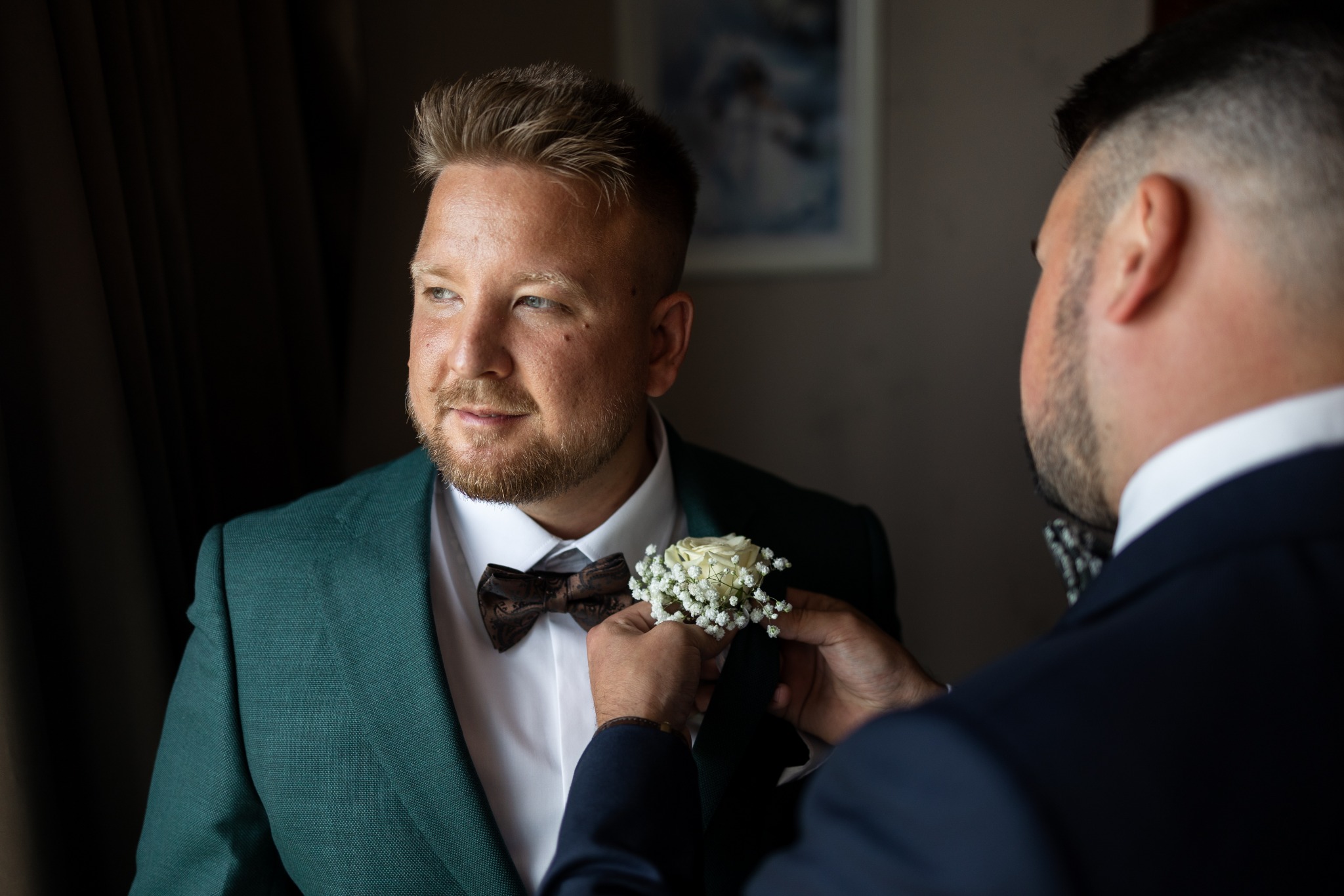 Pan młody w szmaragdowym garniturze i brązowej muszce, w Warszawie, poprawia kwiat w butonierce, spoglądając w lewo.