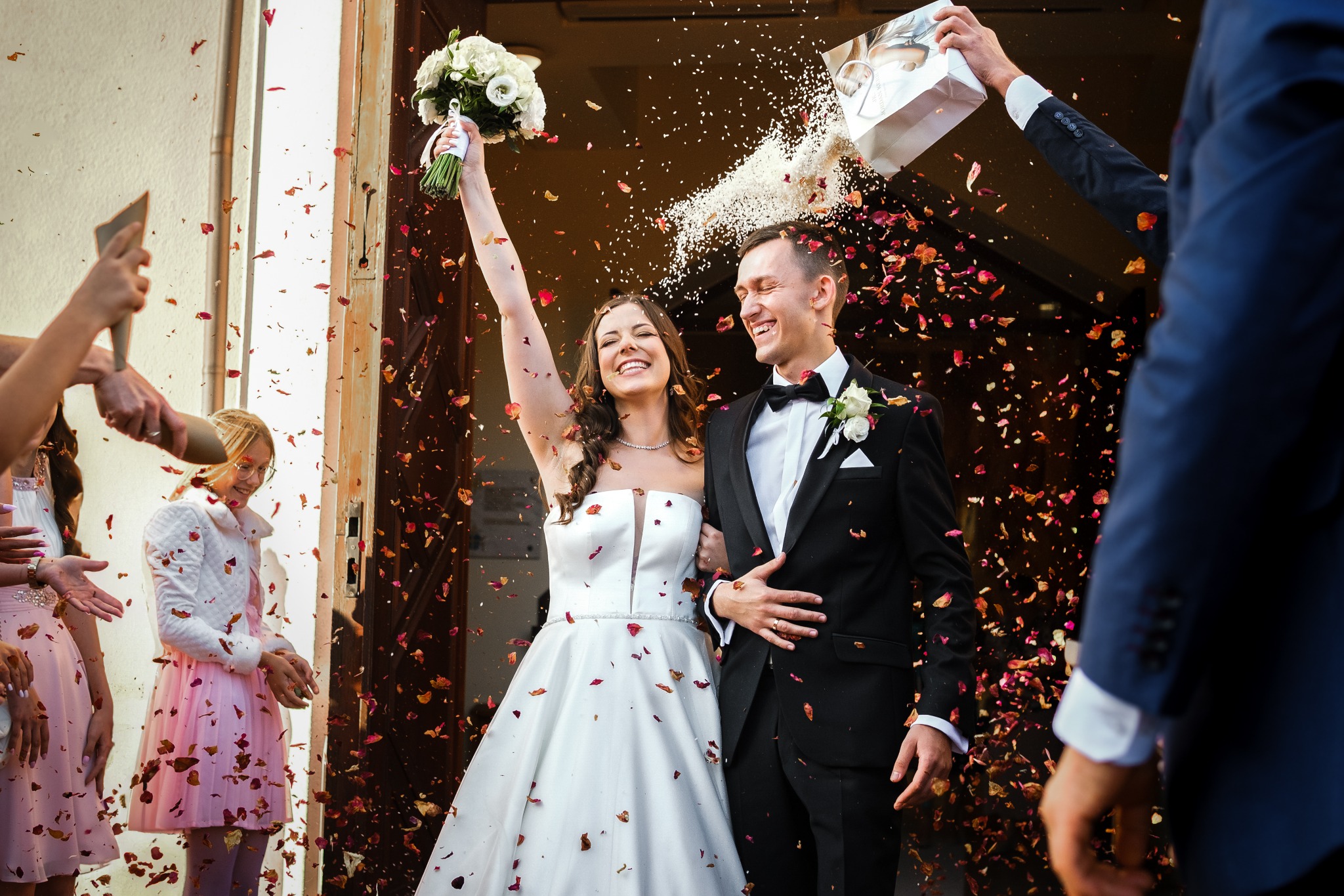 Uśmiechnięta para młoda wychodzi z kościoła, obsypywana płatkami kwiatów i ryżem, panna młoda trzyma bukiet białych róż, goście weselni z telefonami w dłoniach.