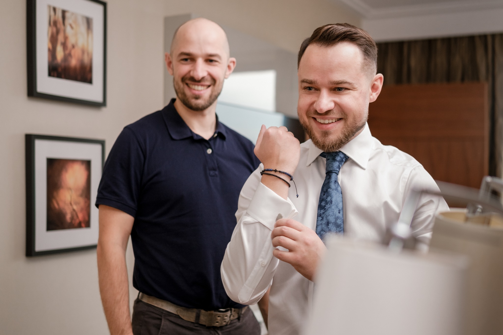 Uśmiechnięty pan młody zapina mankiet w białej koszuli i niebieskim krawacie, za nim stoi drugi mężczyzna w granatowej koszulce polo, w tle obrazy w czarnych ramkach.