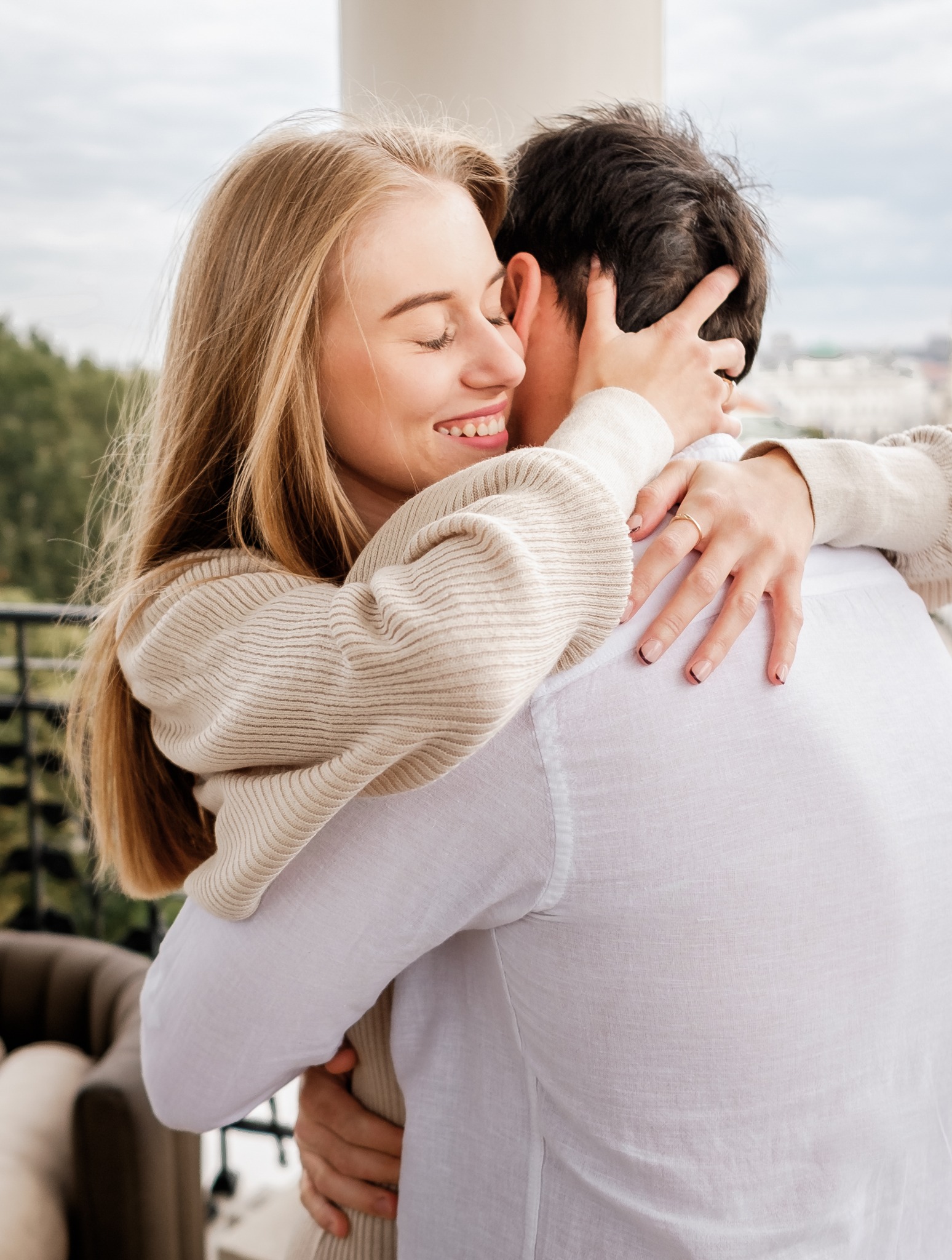 Uśmiechnięta blondynka w beżowym swetrze obejmuje mężczyznę w białej koszuli na tle balkonu z widokiem na miasto, na jej palcu widoczny złoty pierścionek.