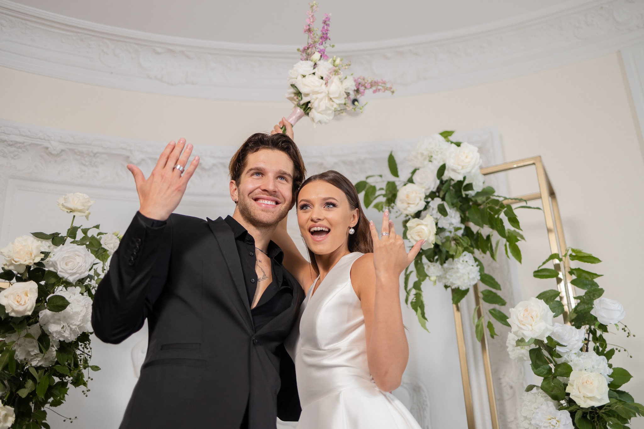 Uśmiechnięta para w strojach ślubnych pozuje z podniesionymi rękami i pierścionkami, panna młoda trzyma bukiet nad głową pana młodego, w tle dekoracja kwiatowa i białe ściany.