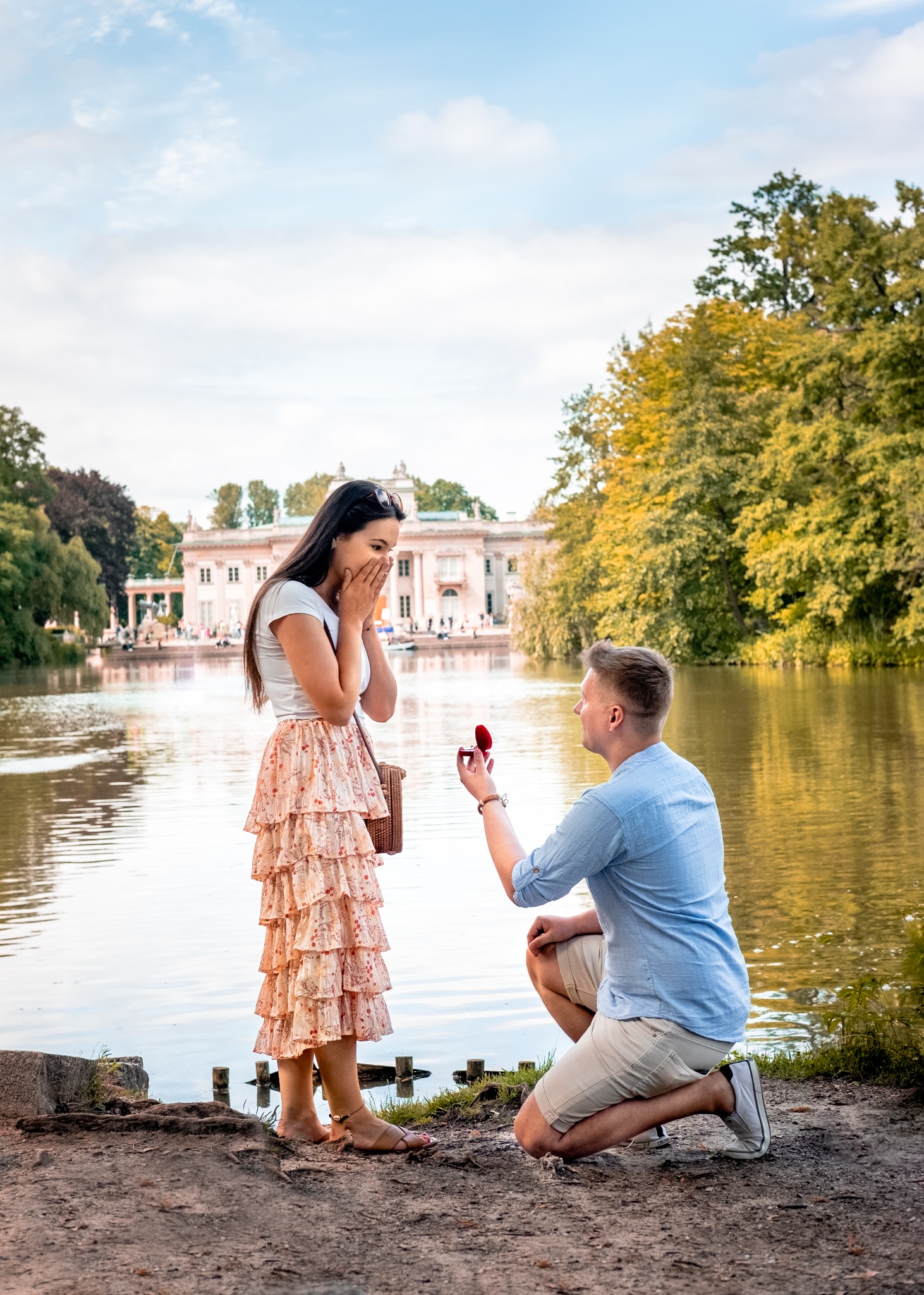 Mężczyzna klęczy przed kobietą nad jeziorem, trzymając otwarte pudełko z pierścionkiem zaręczynowym; w tle Pałac na Wodzie w Łazienkach Królewskich.