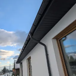 Fragment elewacji domu z widocznym systemem orynnowania w kolorze grafitowym, drewnianą stolarką okienną i świeżo otynkowaną, białą elewacją w zimowej scenerii.