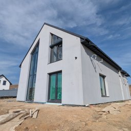 RP. Radosław Paździor - Nowoczesny dom z jasną elewacją i dużymi oknami, widoczny z dołu na tle błękitnego nieba. Teren wokół domu w trakcie prac wykończeniowych.