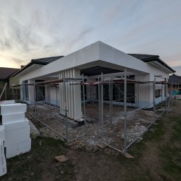 RP. Radosław Paździor - Dom w trakcie prac elewacyjnych w Bachowicach, widoczne rusztowania, częściowo ocieplone ściany i stos styropianu. Ujęcie z dołu, w tle zachmurzone niebo.