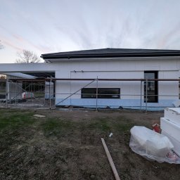 RP. Radosław Paździor - Dom w trakcie ocieplania, widoczne rusztowania i styropian. Prace elewacyjne w toku, jasny styropian na ścianach budynku, błękitna podmurówka.