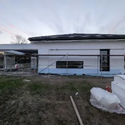 Dom w trakcie ocieplania, widoczne rusztowania i styropian. Prace elewacyjne w toku, jasny styropian na ścianach budynku, błękitna podmurówka.