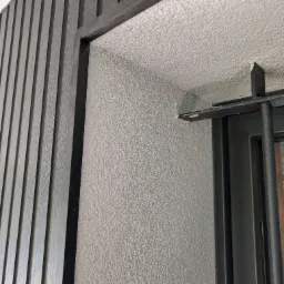 Fragment elewacji z szarymi, drewnopodobnymi panelami i tynkiem strukturalnym wokół okna z czarną ramą. Detal wykończenia ściany zewnętrznej.