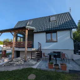 Dom z jasną elewacją, ciemnym dachem i drewnianą pergolą. Widoczne spiralne schody, okna dachowe, zbiornik na wodę i częściowo zasłonięty van. Ujęcie w słoneczny dzień.