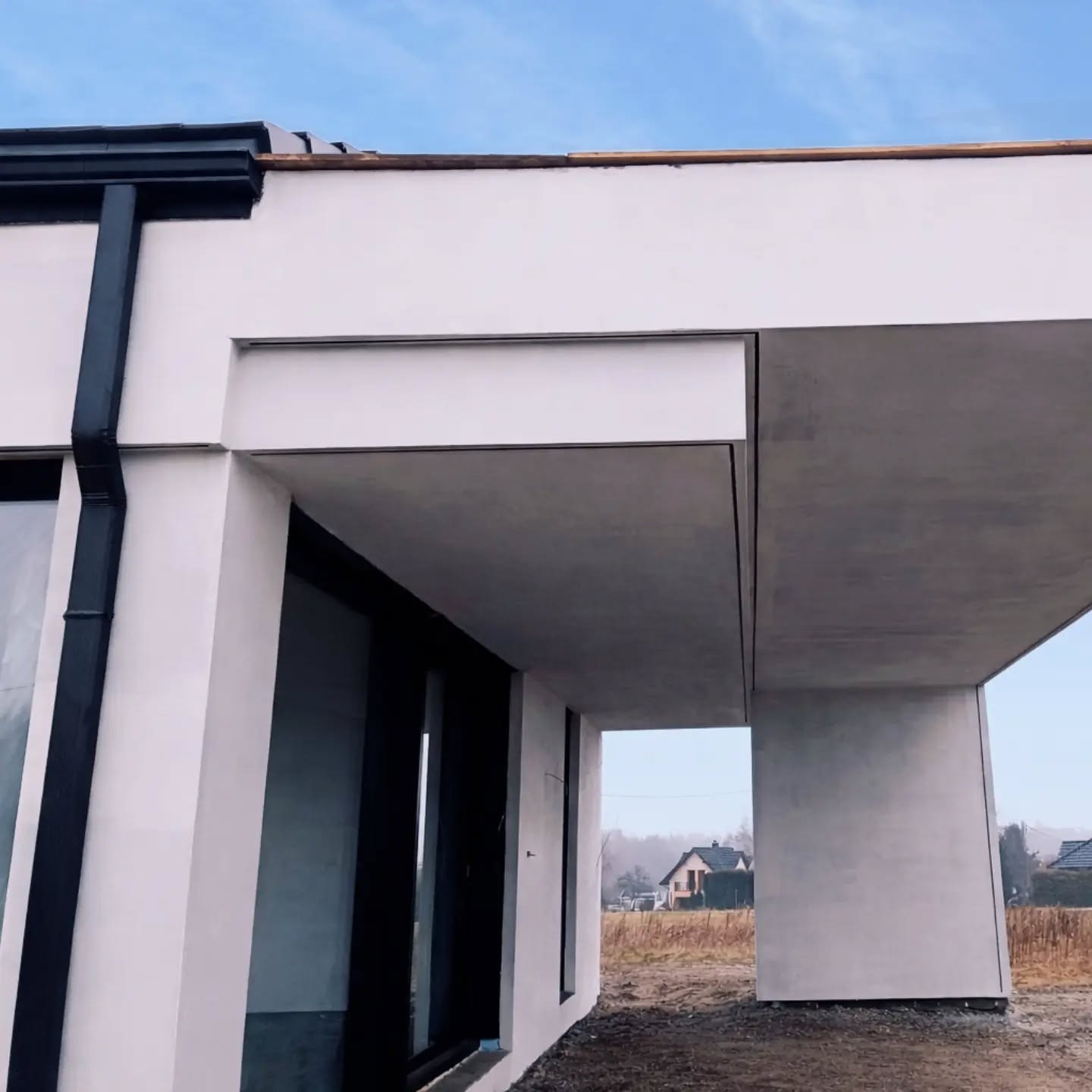 Nowoczesna elewacja domu jednorodzinnego z minimalistycznym wykończeniem, widok z zewnątrz, zadaszenie nad wejściem, czarne rynny, jasna kolorystyka.