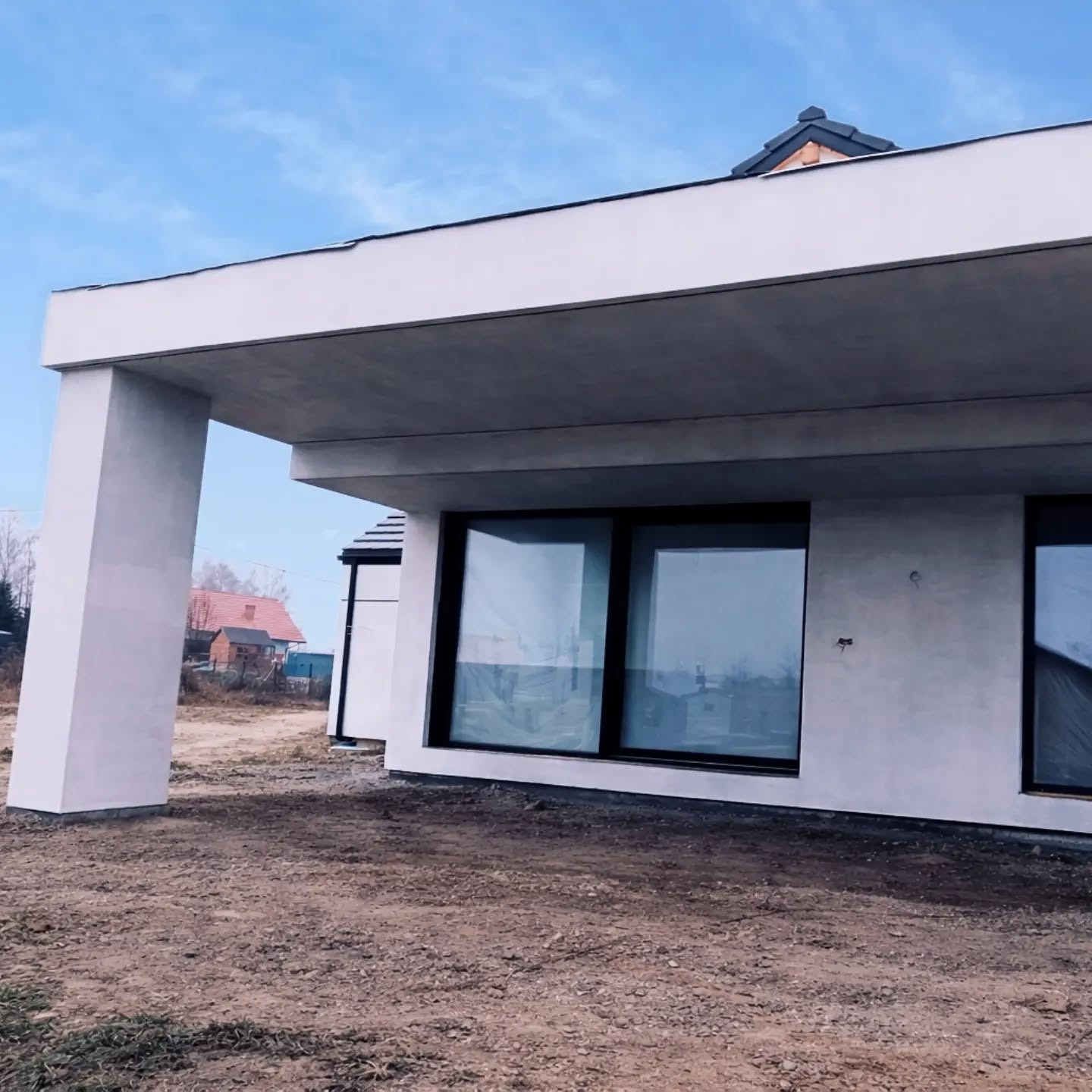 Nowoczesny dom jednorodzinny z płaskim dachem i dużymi oknami, wykończony jasnym tynkiem. Widoczny fragment elewacji z kolumną podtrzymującą zadaszenie.