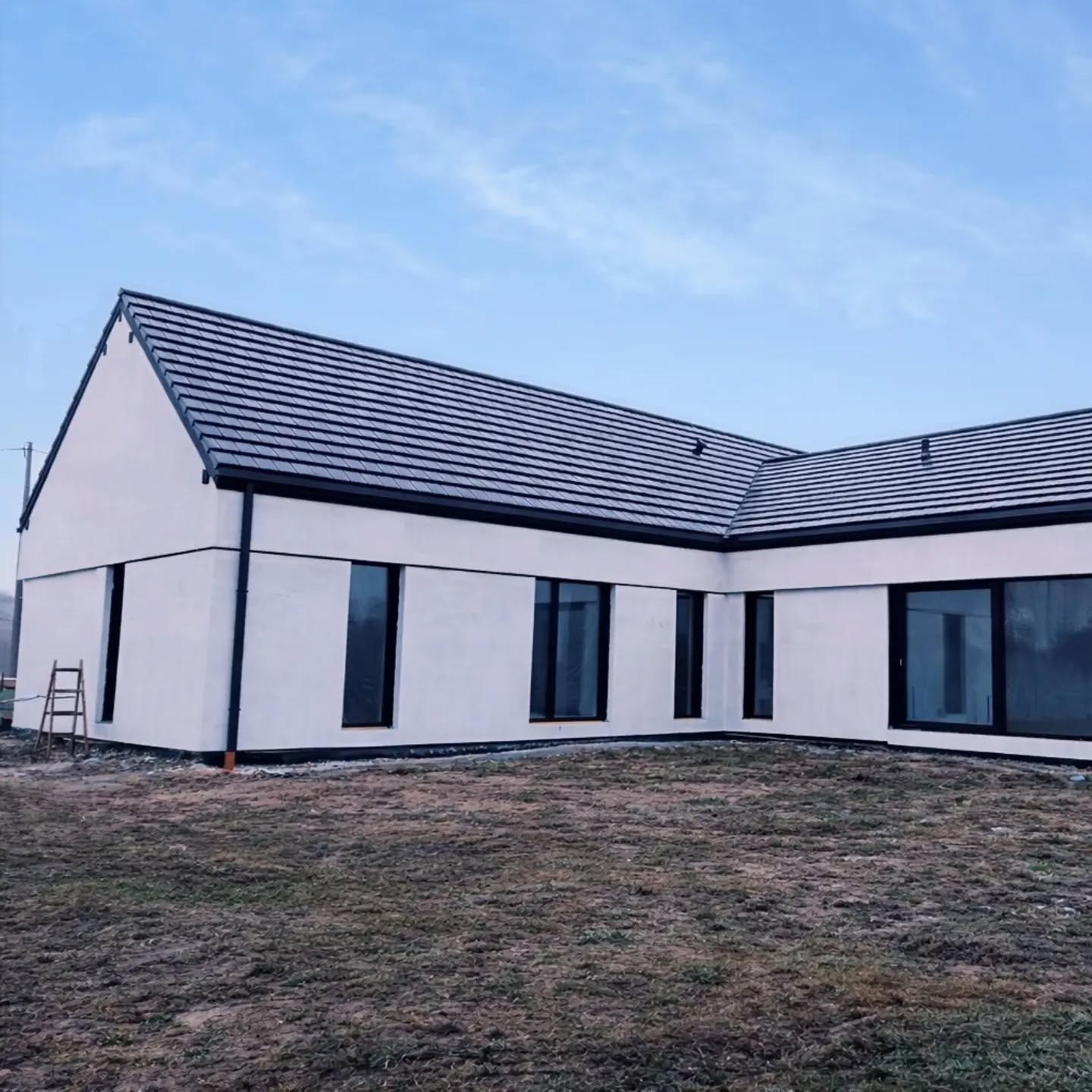 Nowoczesny, parterowy dom z białą elewacją i ciemną stolarką okienną. Widoczny fragment dachu z ciemnej dachówki. Na pierwszym planie trawnik.