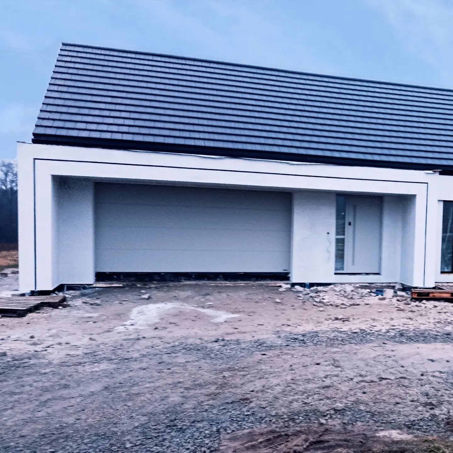 Nowoczesny dom jednorodzinny z płaskim dachem i garażem w bryle, świeżo po pracach elewacyjnych. Widoczne drobne pozostałości po budowie.