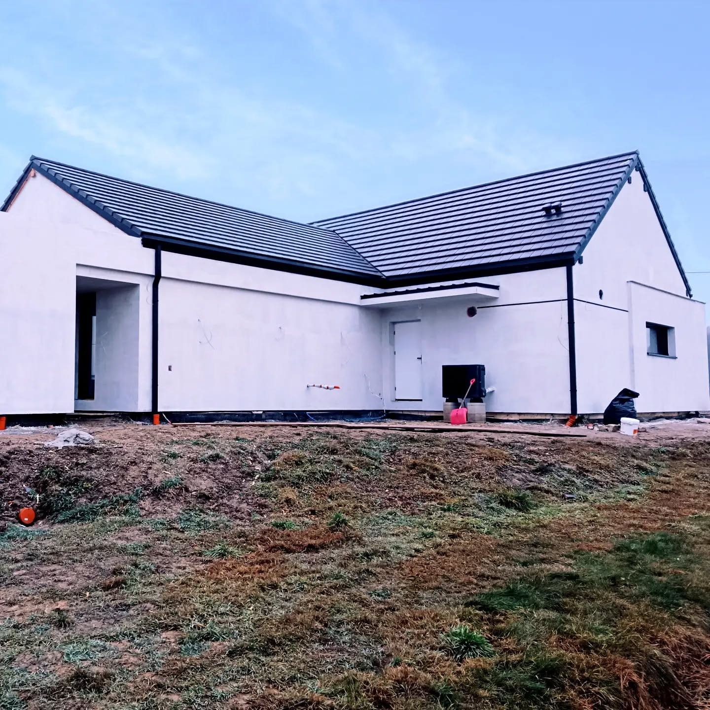 Nowoczesny dom jednorodzinny z białymi ścianami i ciemną dachówką, w trakcie wykończenia elewacji. Widoczne narzędzia i materiały budowlane na posesji.