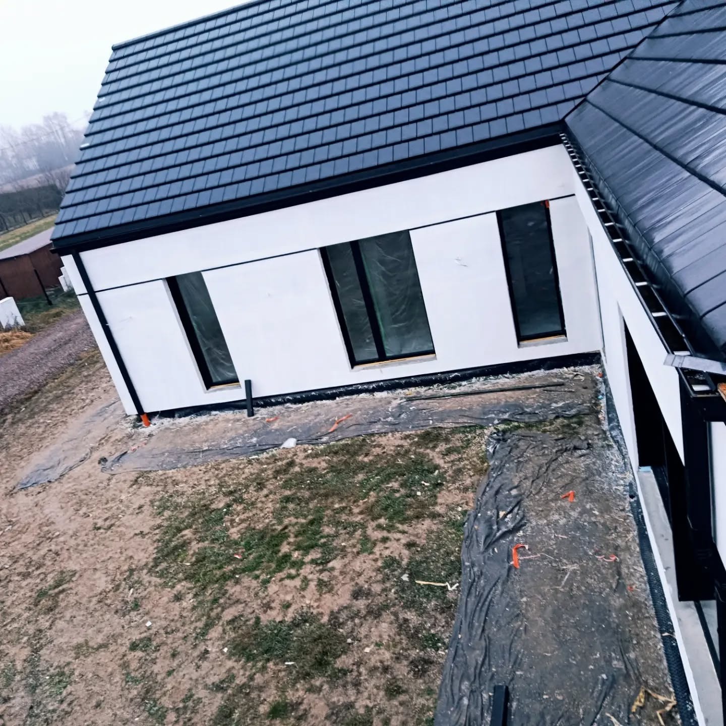 Nowoczesny dom z białą elewacją i grafitowym dachem, widok z góry. Okna zabezpieczone folią, teren wokół przygotowany pod dalsze prace.