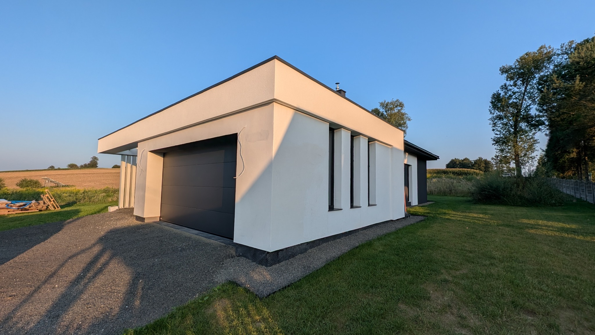 Nowoczesny dom jednorodzinny z garażem, wykończony białą elewacją i grafitową bramą garażową, otoczony zielonym trawnikiem i polami w tle.