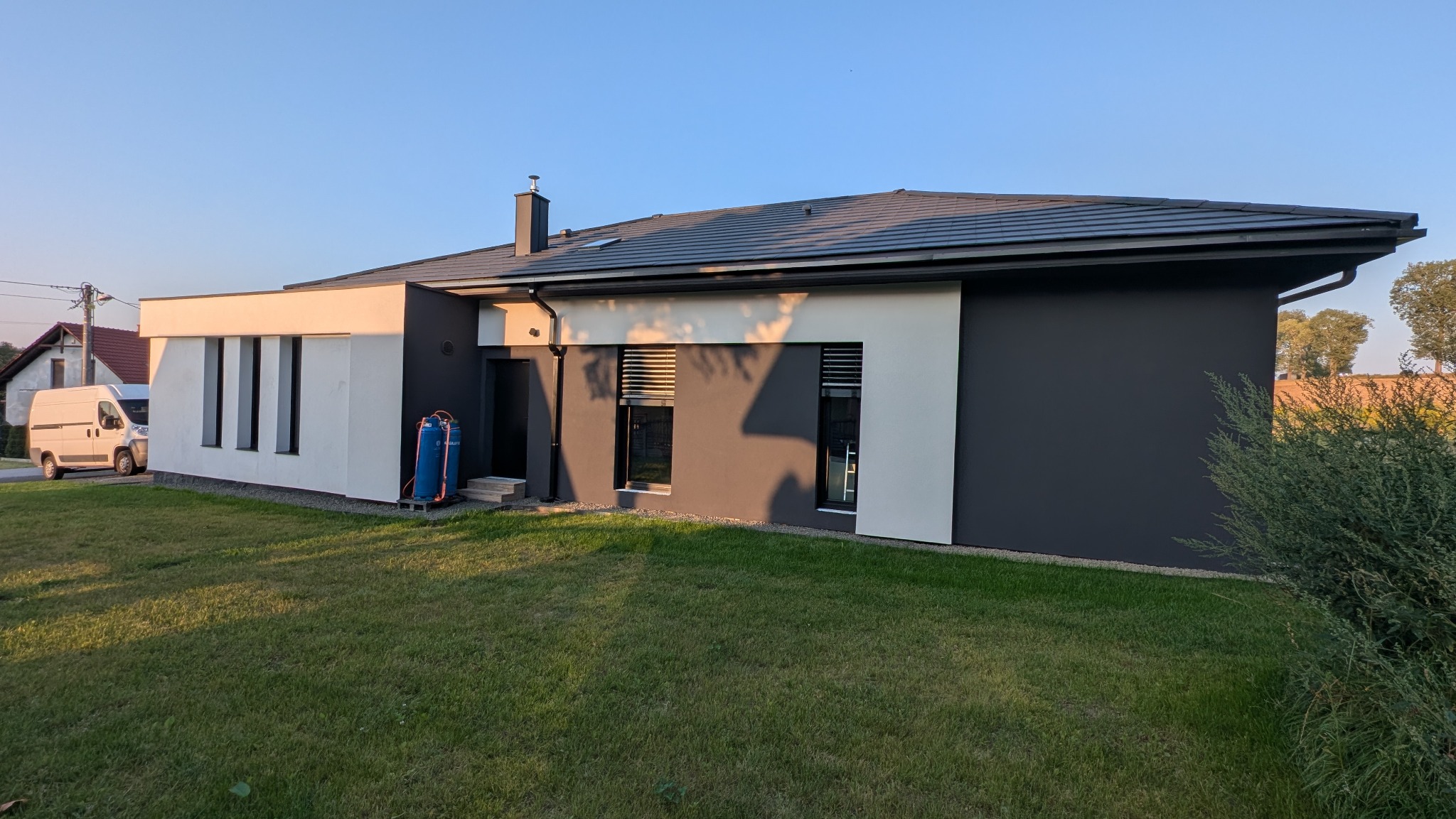 Nowoczesny dom jednorodzinny z grafitową elewacją i białymi akcentami, widok z ogrodu w słoneczny dzień. Na trawniku stoi niebieski sprzęt malarski.