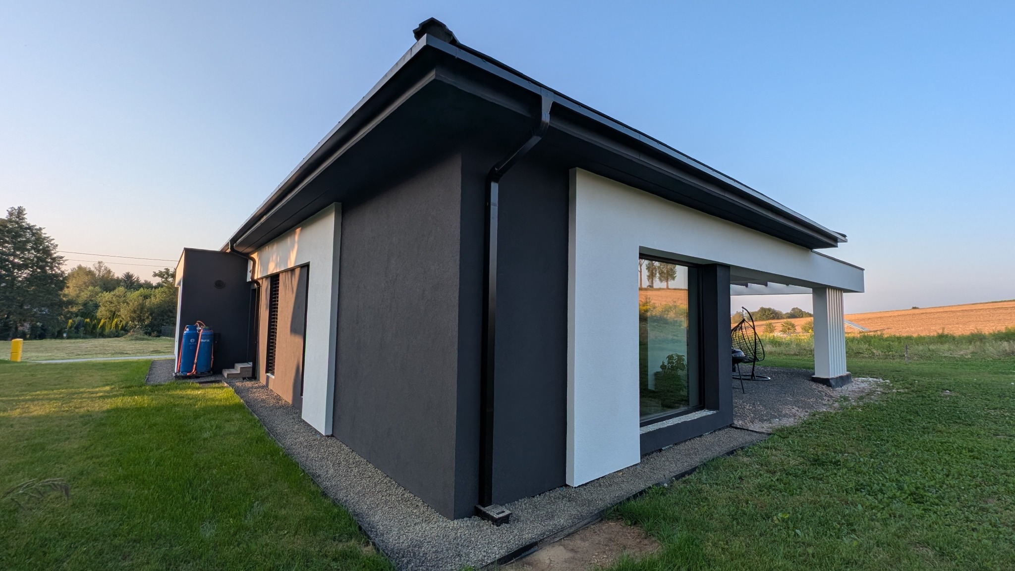 Nowoczesny dom jednorodzinny z elewacją w odcieniach szarości i bieli, widok z zewnątrz, zieleń trawnika i pola w tle, minimalistyczny design.