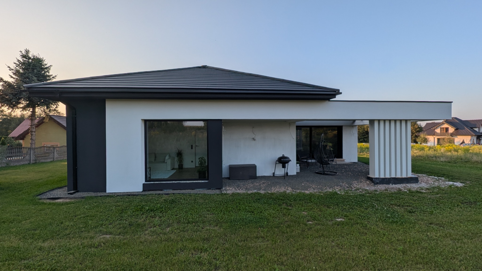 Nowoczesny dom jednorodzinny z płaskim dachem i elewacją w kolorach bieli i grafitu. Widoczne duże okna i taras z meblami ogrodowymi. Trawa na pierwszym planie.