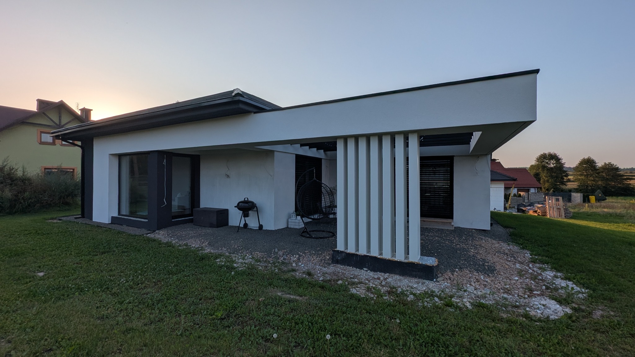 Nowoczesny dom jednorodzinny z białymi ścianami i elementami dekoracyjnymi z pionowych listew. Widoczne detale wykończenia elewacji i otoczenie domu.