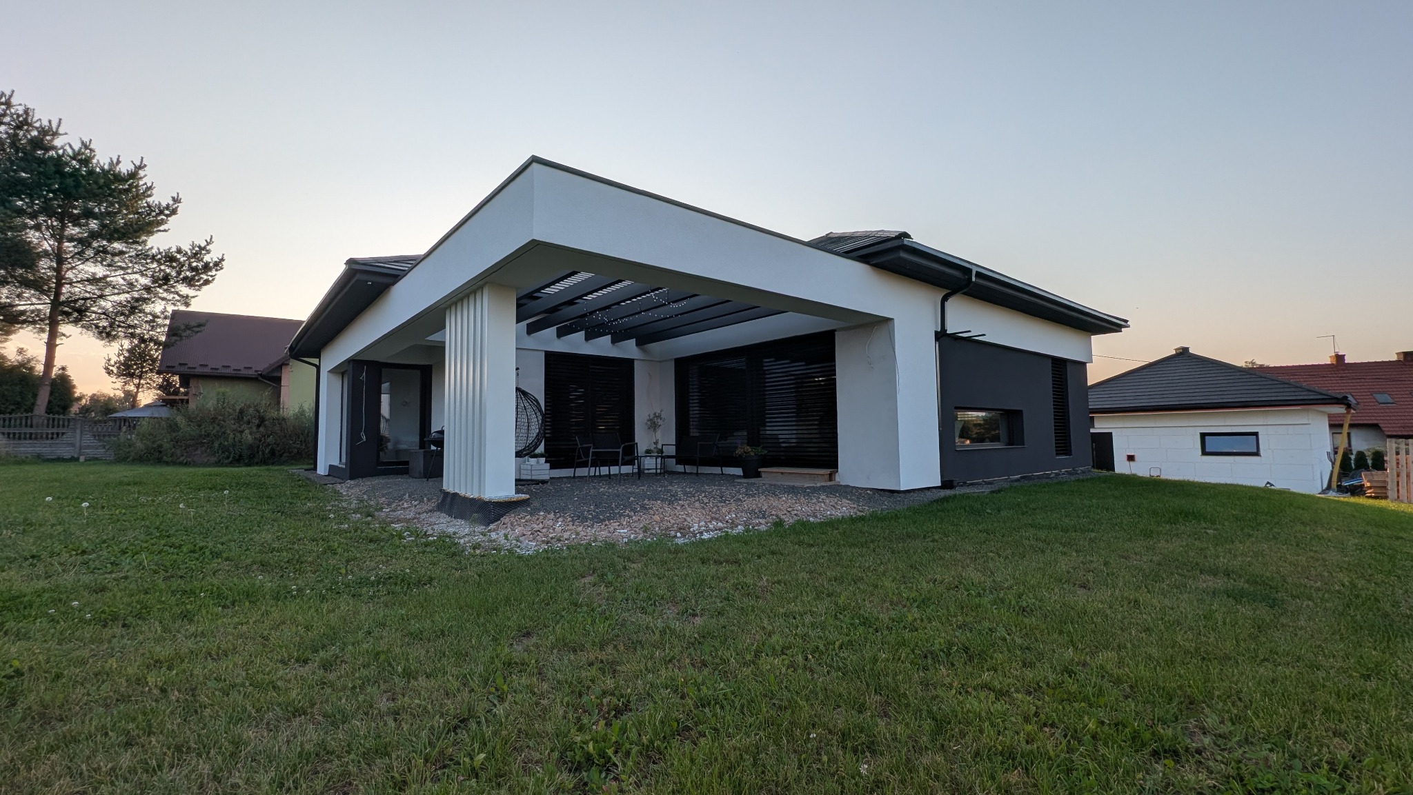 Nowoczesny dom jednorodzinny z płaskim dachem i pergolą nad tarasem, wykończony w bieli i szarości, otoczony zielonym trawnikiem. Widok z poziomu gruntu.