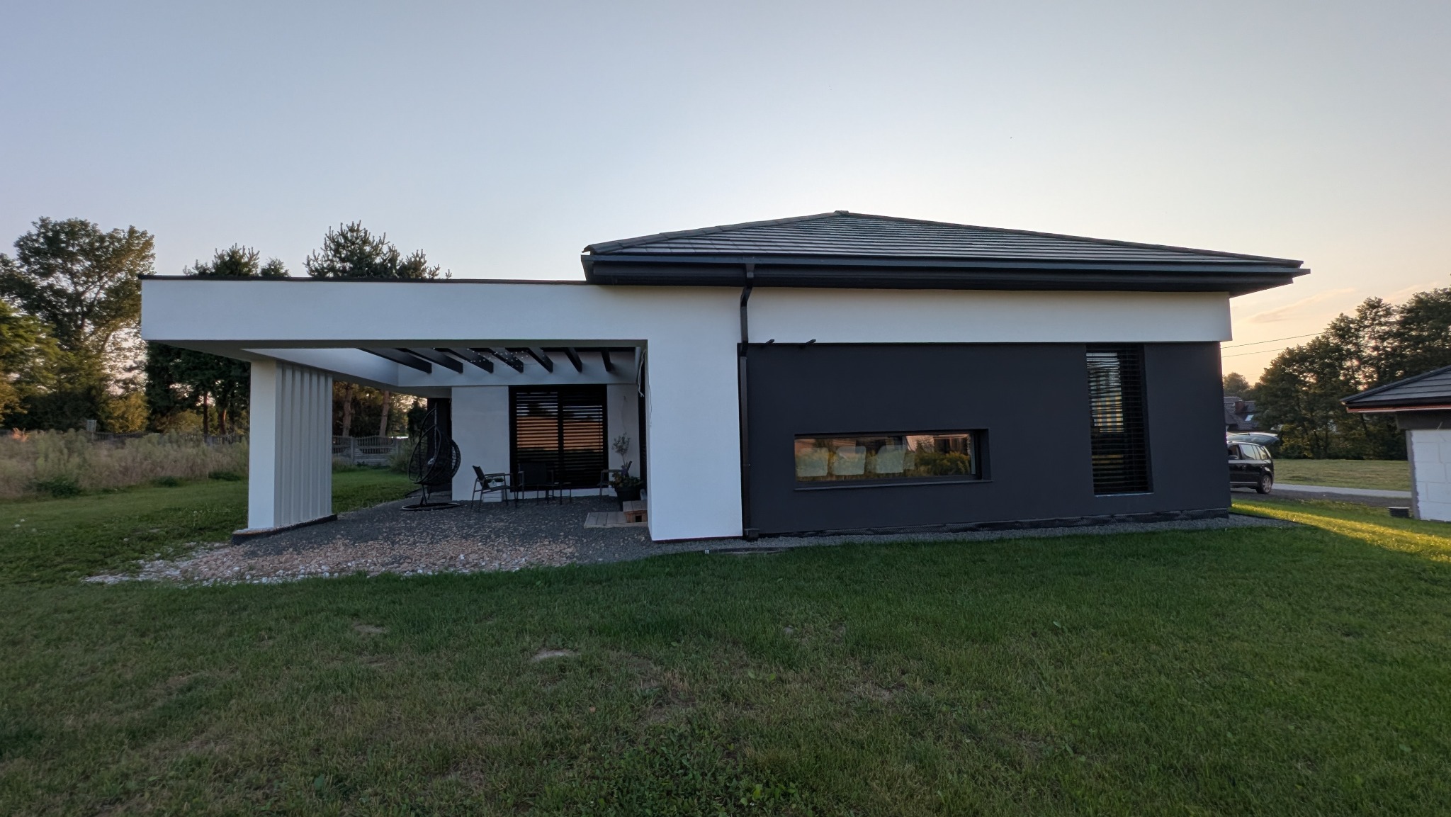 Nowoczesny dom jednorodzinny z płaskim dachem i minimalistyczną elewacją w odcieniach bieli i grafitu, z zadaszonym tarasem i starannie zagospodarowanym otoczeniem.