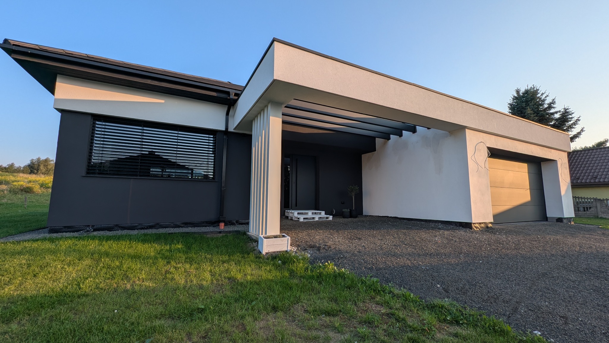 Nowoczesny dom jednorodzinny z grafitową elewacją i białymi akcentami, widok z zewnątrz, w tle zieleń. Minimalistyczny design z garażem i zadaszonym wejściem.