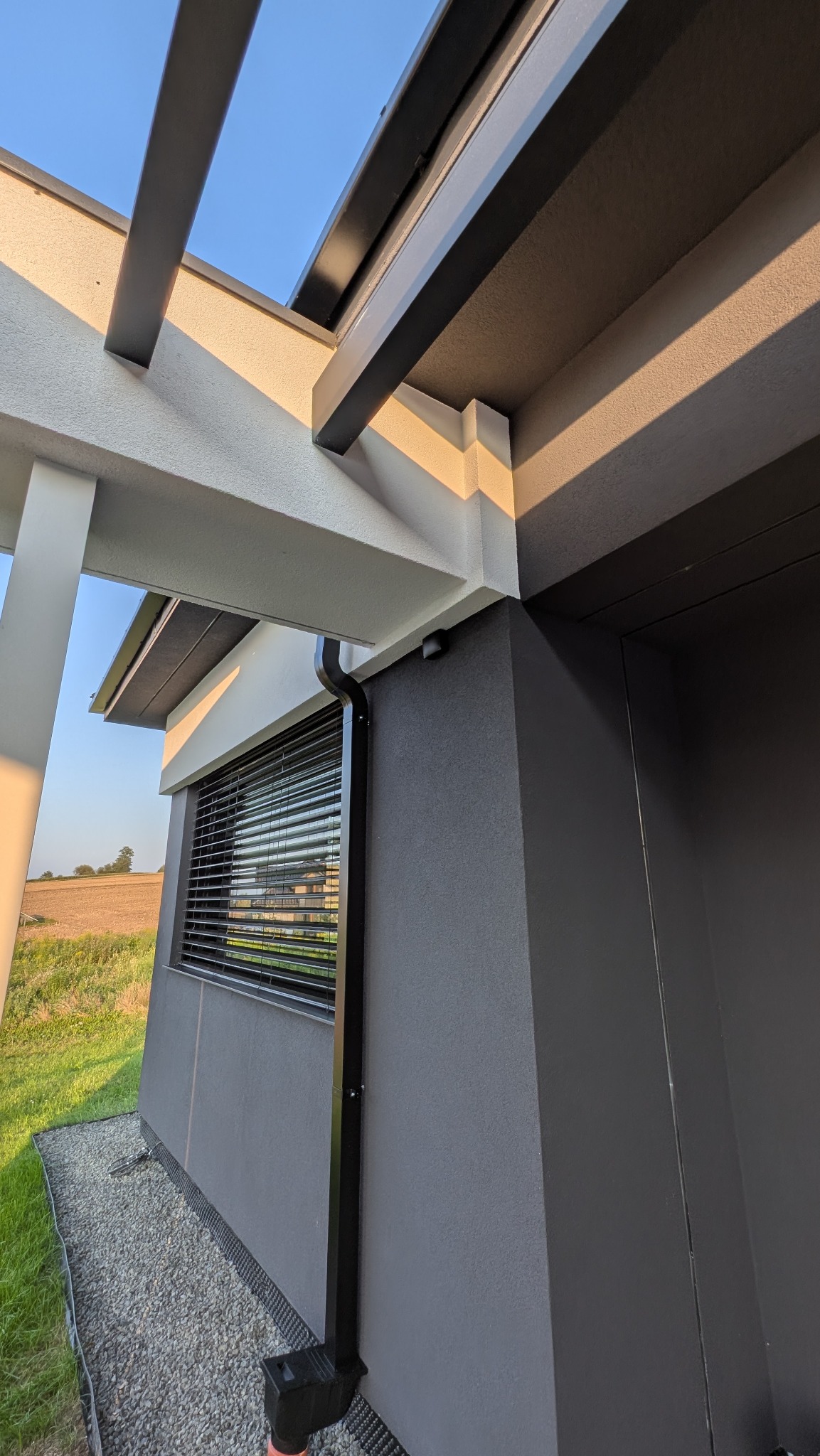Fragment elewacji domu z szarą, teksturowaną ścianą, czarną rynną i oknem z żaluzjami. Widoczne elementy konstrukcyjne pergoli i fragment krajobrazu w tle.