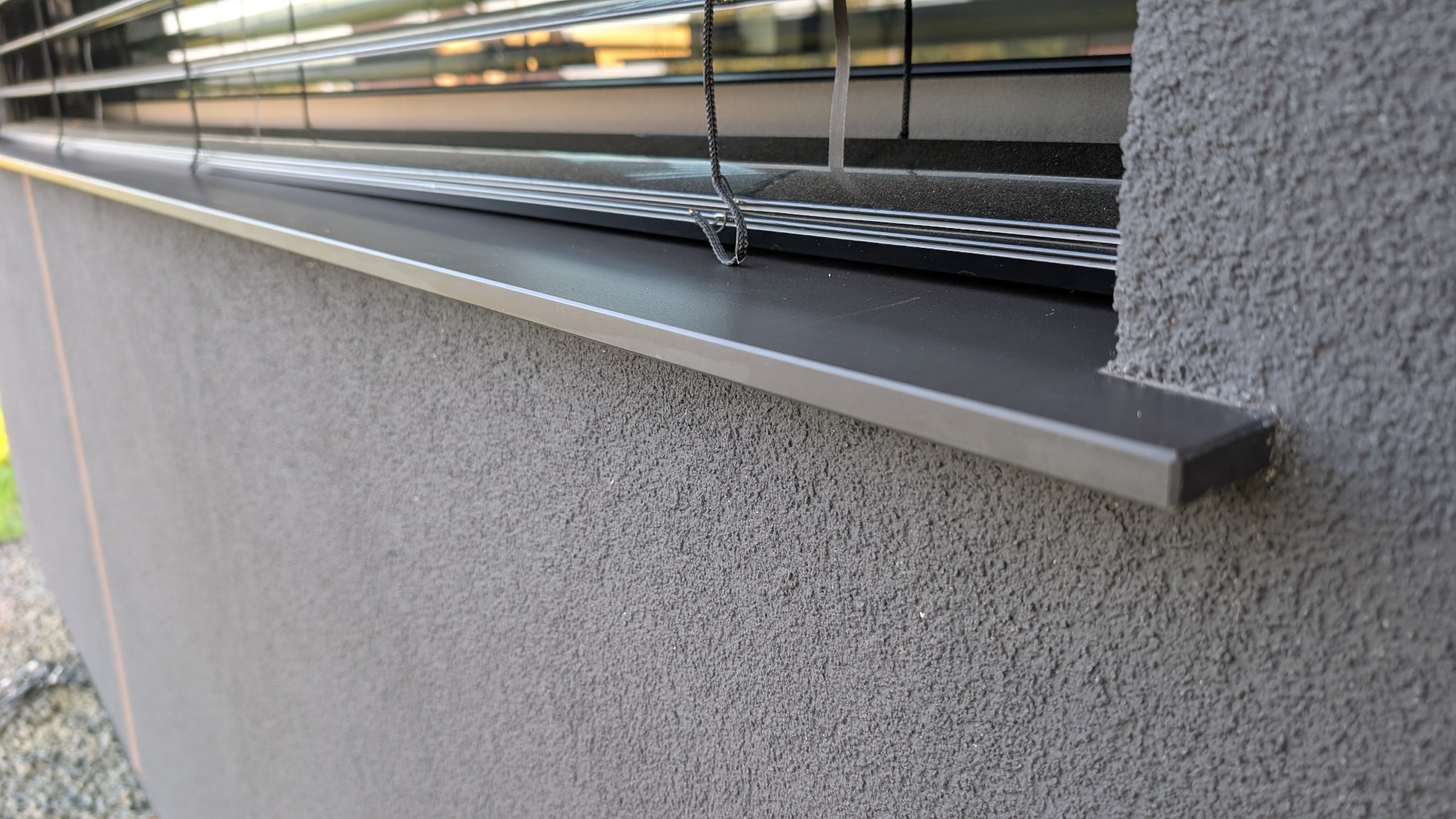 Szara, chropowata elewacja z oknem i metalowym parapetem. Widoczne żaluzje okienne. Detal wykończenia elewacji w nowoczesnym stylu.