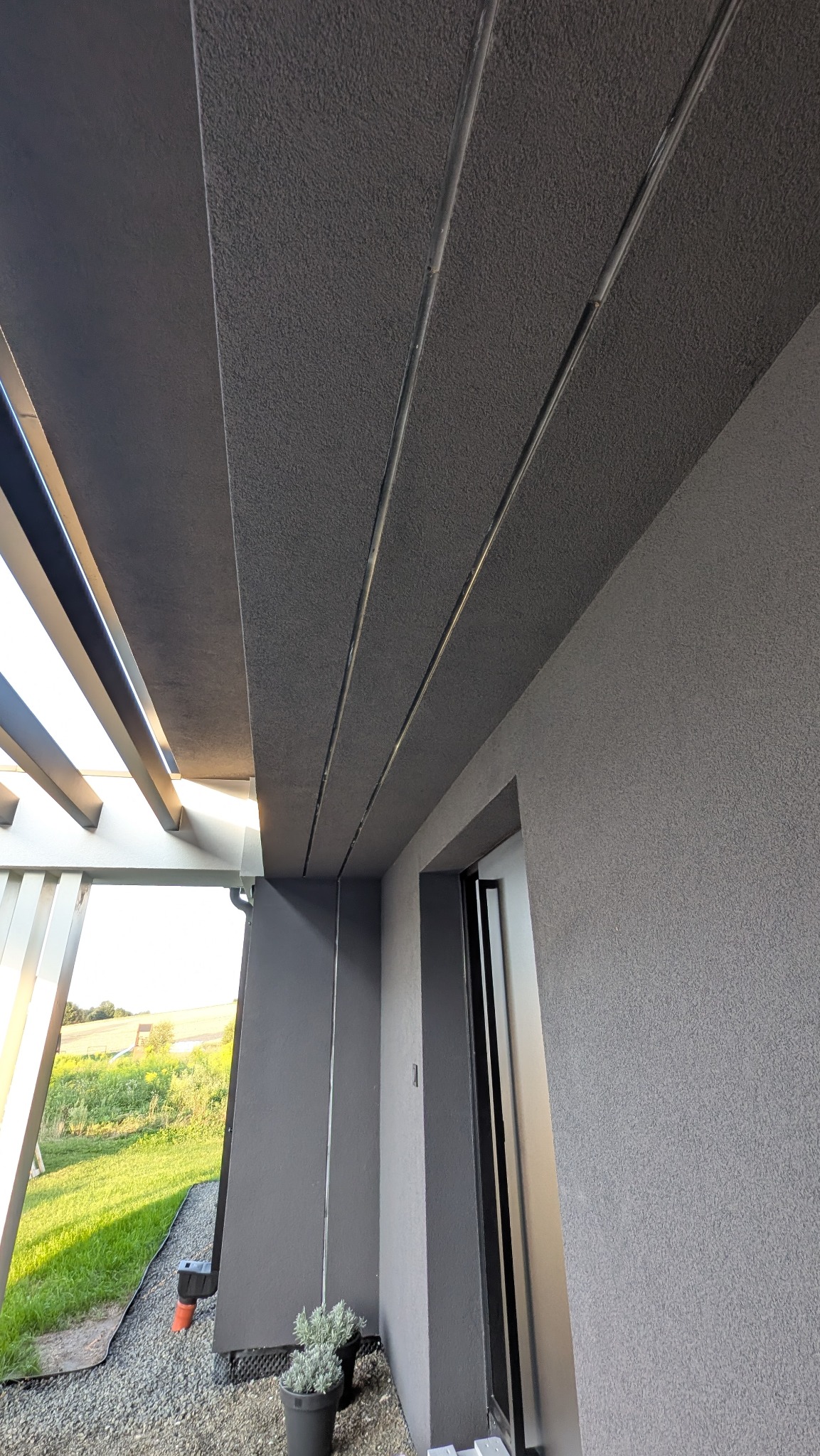 Szara elewacja domu z widoczną strukturą tynku, fragmentem pergoli i rośliną w doniczce. Ujęcie z dołu podkreśla nowoczesny design i precyzję wykonania.