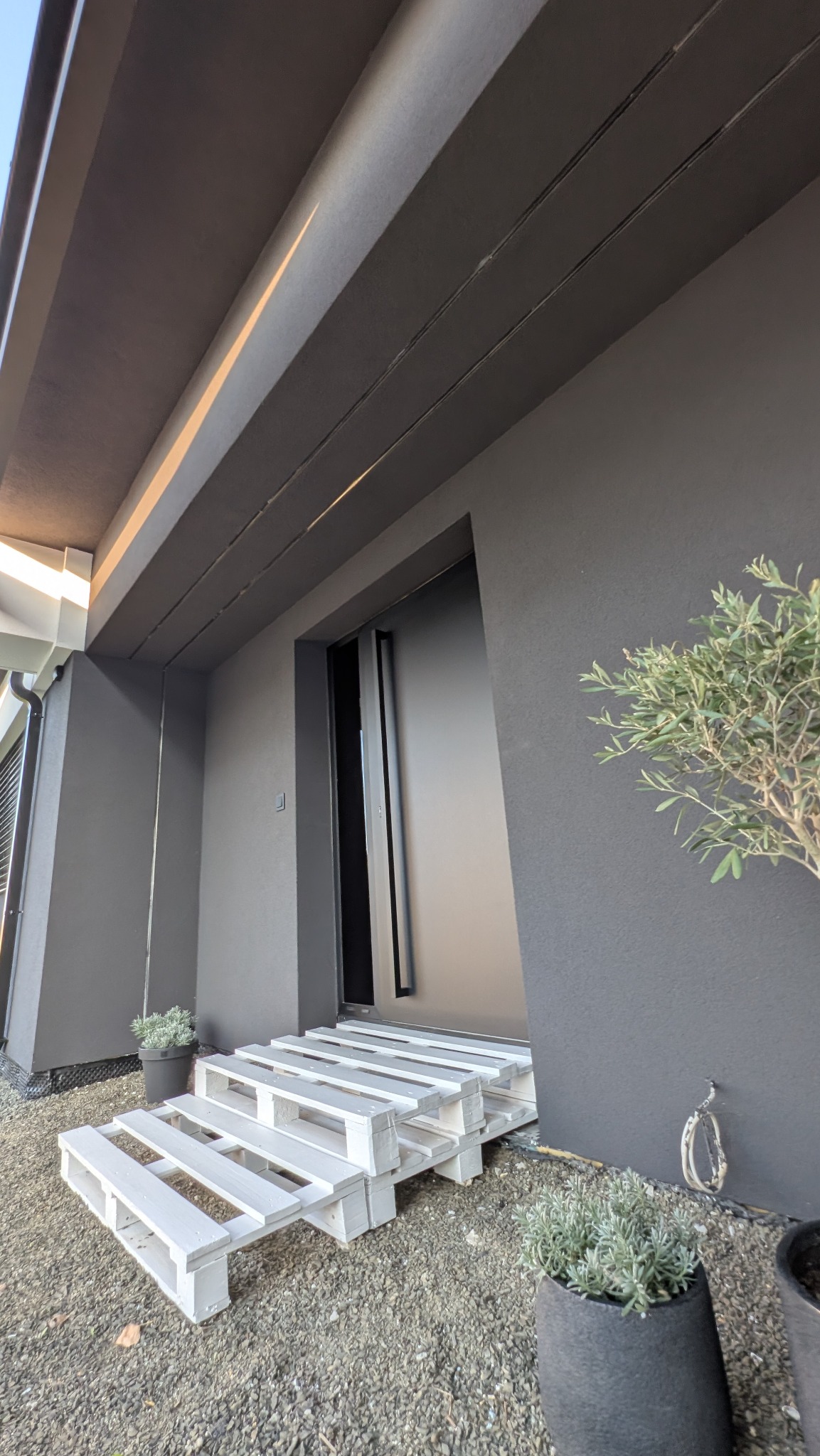 Nowoczesny dom z ciemną elewacją i białymi paletami jako schodami wejściowymi. Minimalistyczny design, rośliny w donicach. Ujęcie z dołu, podkreślające geometrię budynku.