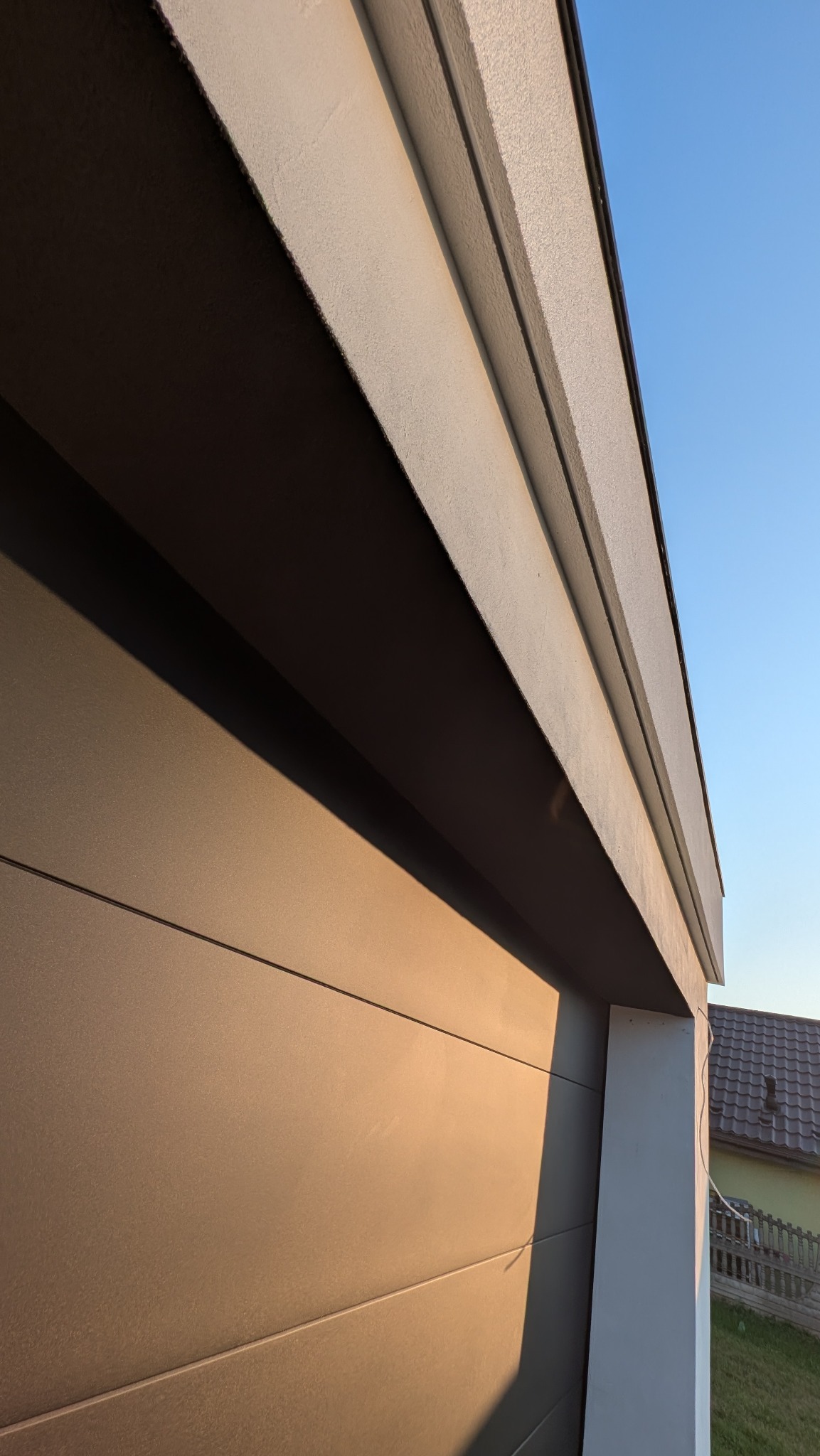 Fragment elewacji budynku z widoczną strukturą tynku i panelami w odcieniach szarości, uchwycony pod kątem z fragmentem nieba w tle.