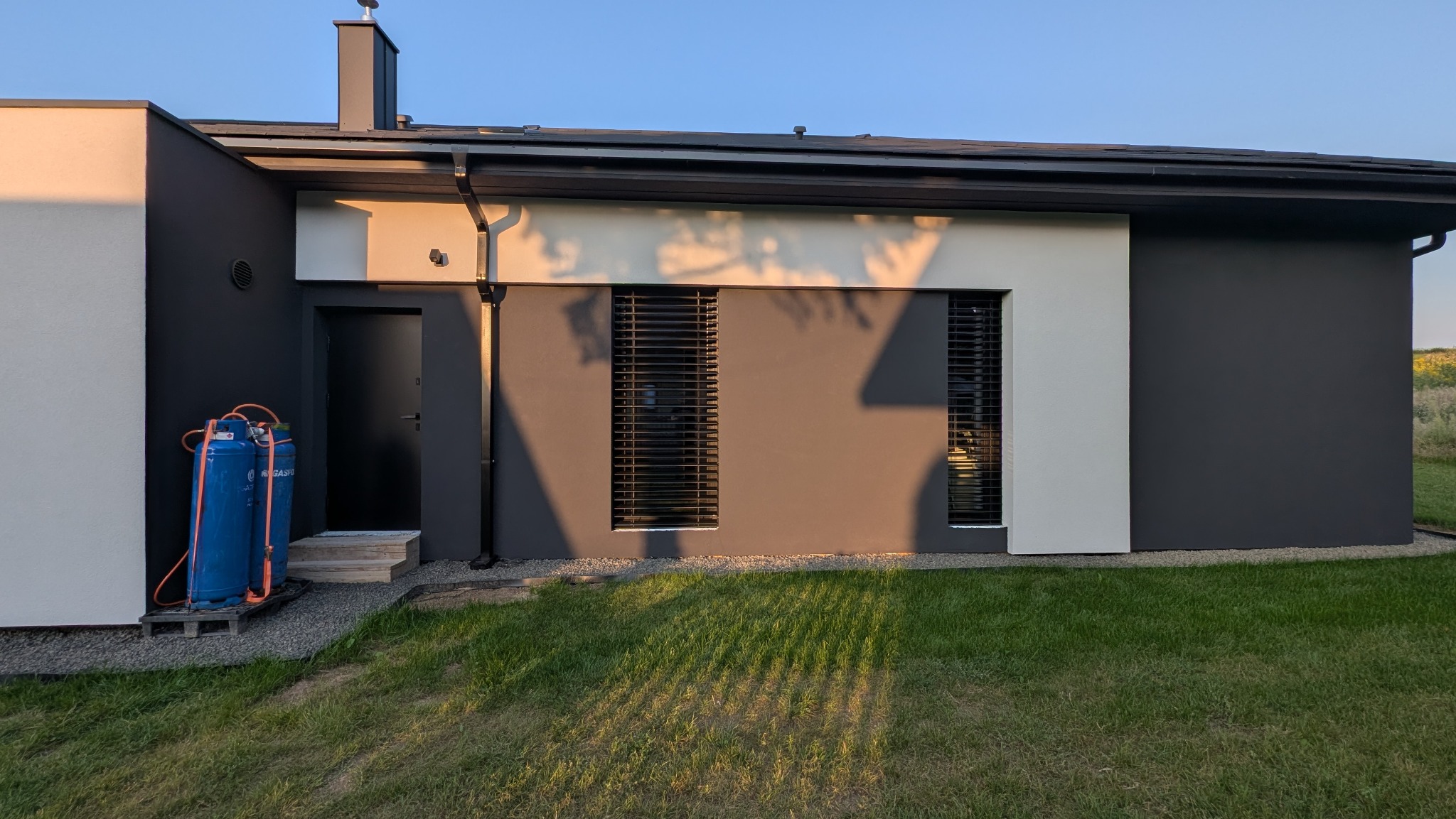 Nowoczesny dom jednorodzinny w Bachowicach z grafitową elewacją i białymi akcentami, widok z zewnątrz, zieleń trawnika i błękitne butle gazowe przy wejściu.