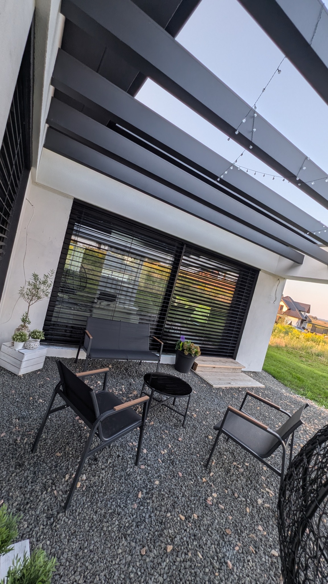 Nowoczesny taras z pergolą i meblami ogrodowymi. Widoczne czarne żaluzje fasadowe, kamienny podkład i oświetlenie dekoracyjne. Minimalistyczny design.