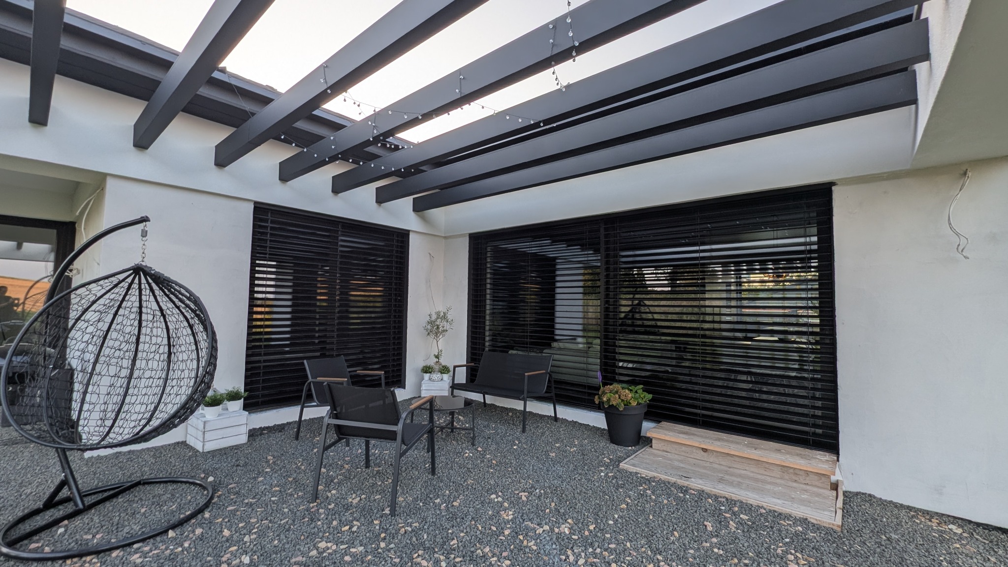 Nowoczesny taras z zadaszeniem z belek, czarnymi żaluzjami okiennymi i szarym żwirowym podłożem. W tle widoczne krzesła i rośliny doniczkowe.
