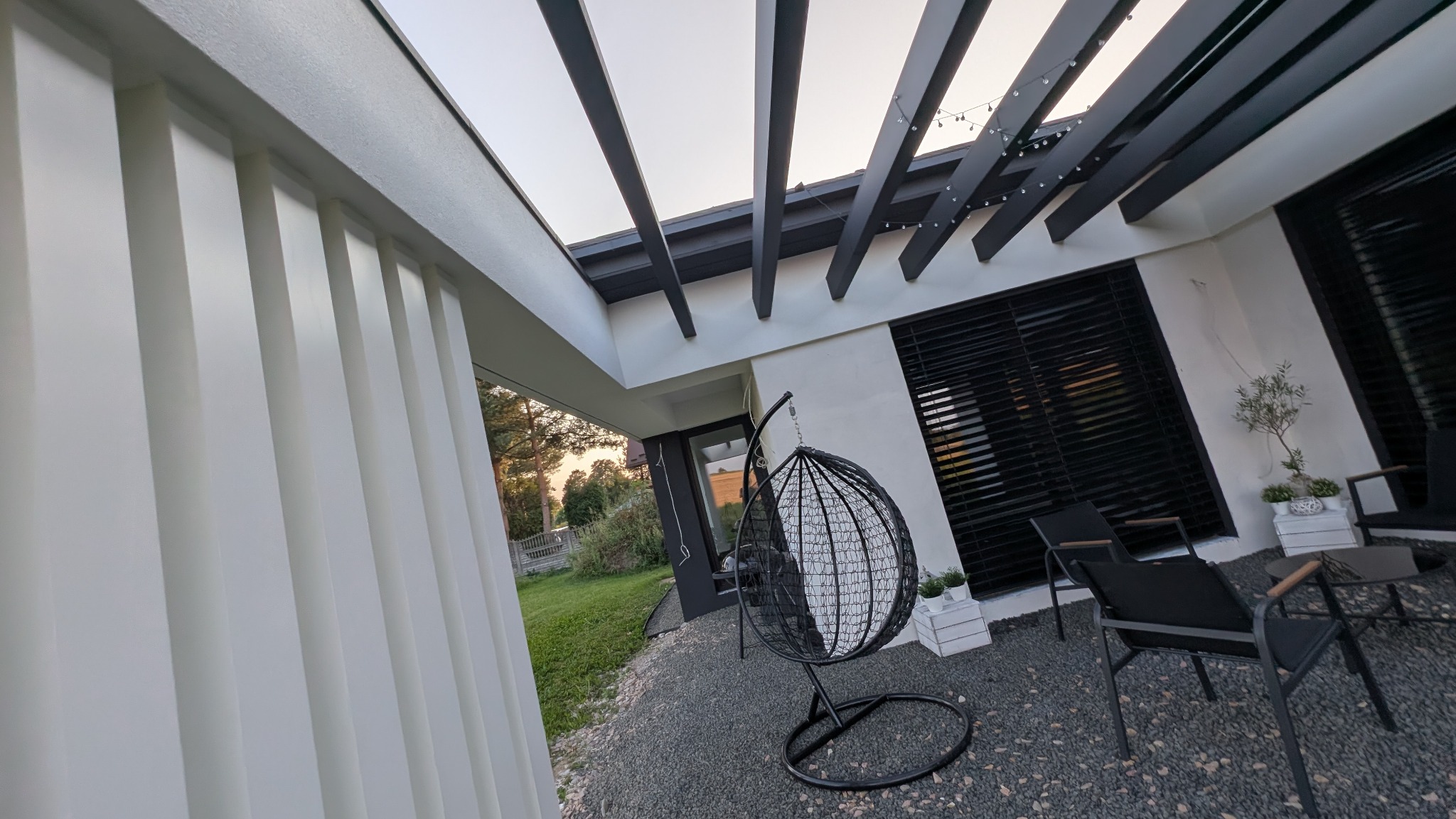 Nowoczesna elewacja domu z pergolą i wiszącym fotelem. Białe ściany kontrastują z czarnymi elementami. Widok na ogród i zachodzące słońce.
