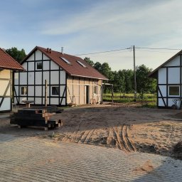 KULTURA ZIELENI SPÓŁKA Z OGRANICZONĄ ODPOWIEDZIALNOŚCIĄ - Trzy budynki w stylu szachulcowym z czerwoną dachówką, widoczne ślady opon na piaszczystym podłożu, przygotowane materiały budowlane i kostka brukowa na pierwszym planie.