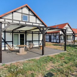 KULTURA ZIELENI SPÓŁKA Z OGRANICZONĄ ODPOWIEDZIALNOŚCIĄ - Nowoczesne domki w Nekli z drewnianą pergolą i tarasem, otoczone dziką roślinnością, widok z zewnątrz w słoneczny dzień.