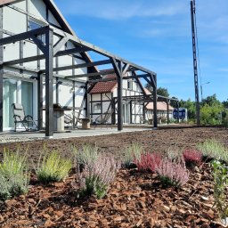 KULTURA ZIELENI SPÓŁKA Z OGRANICZONĄ ODPOWIEDZIALNOŚCIĄ - Nowo założony ogród przydomowy z rabatami wrzosów w odcieniach zieleni, różu i czerwieni, wyściółkowanymi korą, na tle domu w stylu szachulcowym z drewnianą pergolą.
