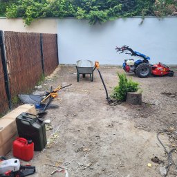 KULTURA ZIELENI SPÓŁKA Z OGRANICZONĄ ODPOWIEDZIALNOŚCIĄ - Teren po wycince drzewa z widocznym pniem, kosiarką rotacyjną, taczką, kanistrami, pilarką, grabiami i łopatą, otoczony płotem z wikliny i białym murem.