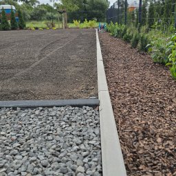 KULTURA ZIELENI SPÓŁKA Z OGRANICZONĄ ODPOWIEDZIALNOŚCIĄ - Przygotowany teren pod ogród z ciemną ziemią, oddzielony betonowym obrzeżem od ścieżki z szarego żwiru i rabaty z korą, z młodym drzewkiem i ogrodzeniem w tle.