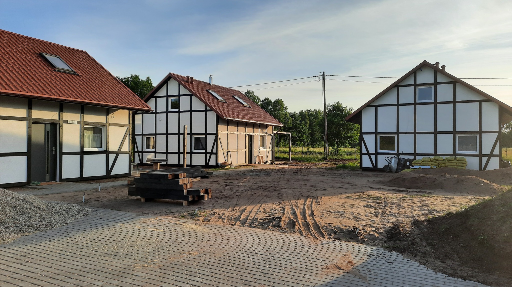 Trzy budynki w stylu szachulcowym z czerwoną dachówką, widoczne ślady opon na piaszczystym podłożu, przygotowane materiały budowlane i kostka brukowa na pierwszym planie.