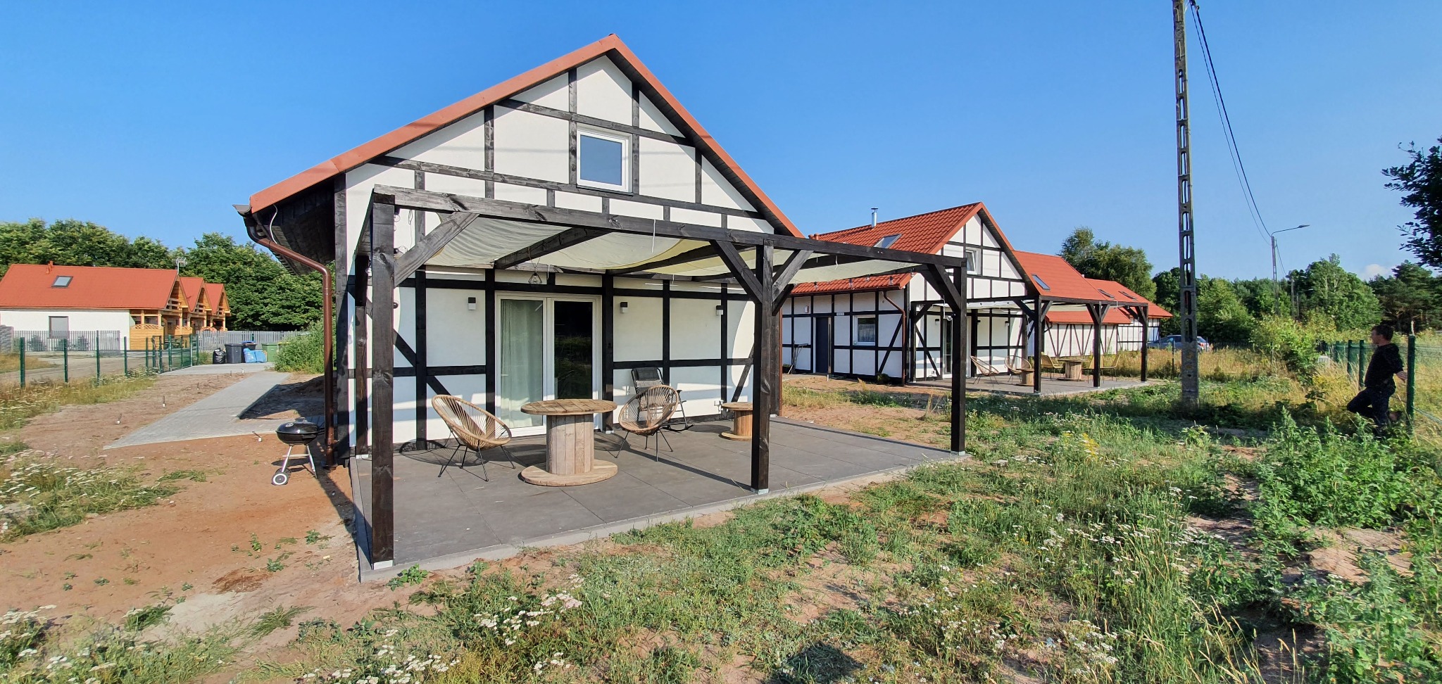 Nowoczesne domki w Nekli z drewnianą pergolą i tarasem, otoczone dziką roślinnością, widok z zewnątrz w słoneczny dzień.