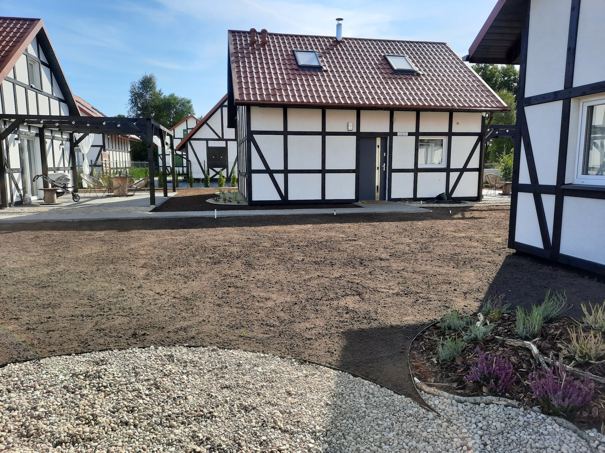 Przygotowany teren pod trawnik przed domami w stylu szachulcowym, z wykończonym obrzeżem z białego kamienia i rabatami z wrzosami.