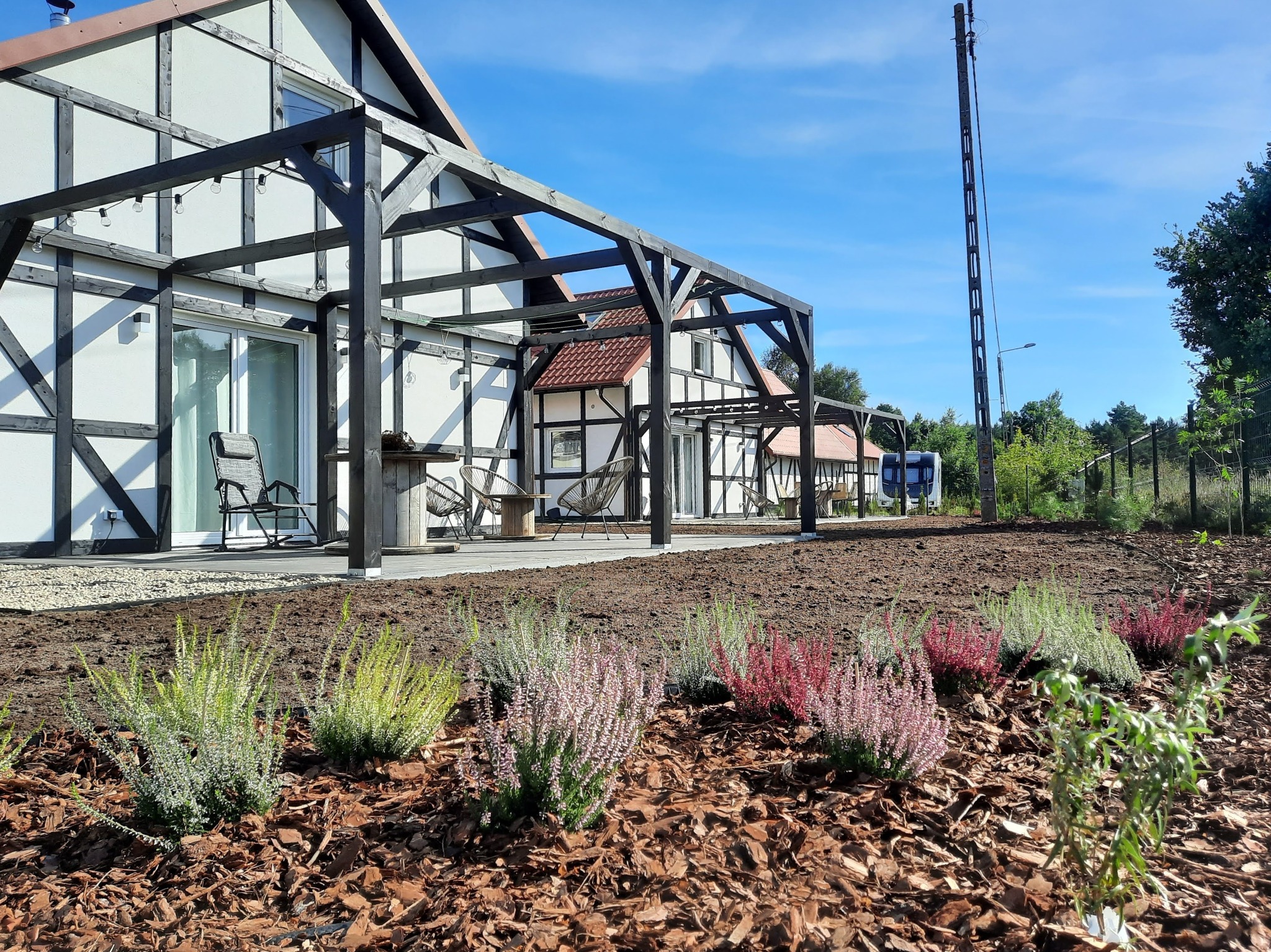 Nowo założony ogród przydomowy z rabatami wrzosów w odcieniach zieleni, różu i czerwieni, wyściółkowanymi korą, na tle domu w stylu szachulcowym z drewnianą pergolą.