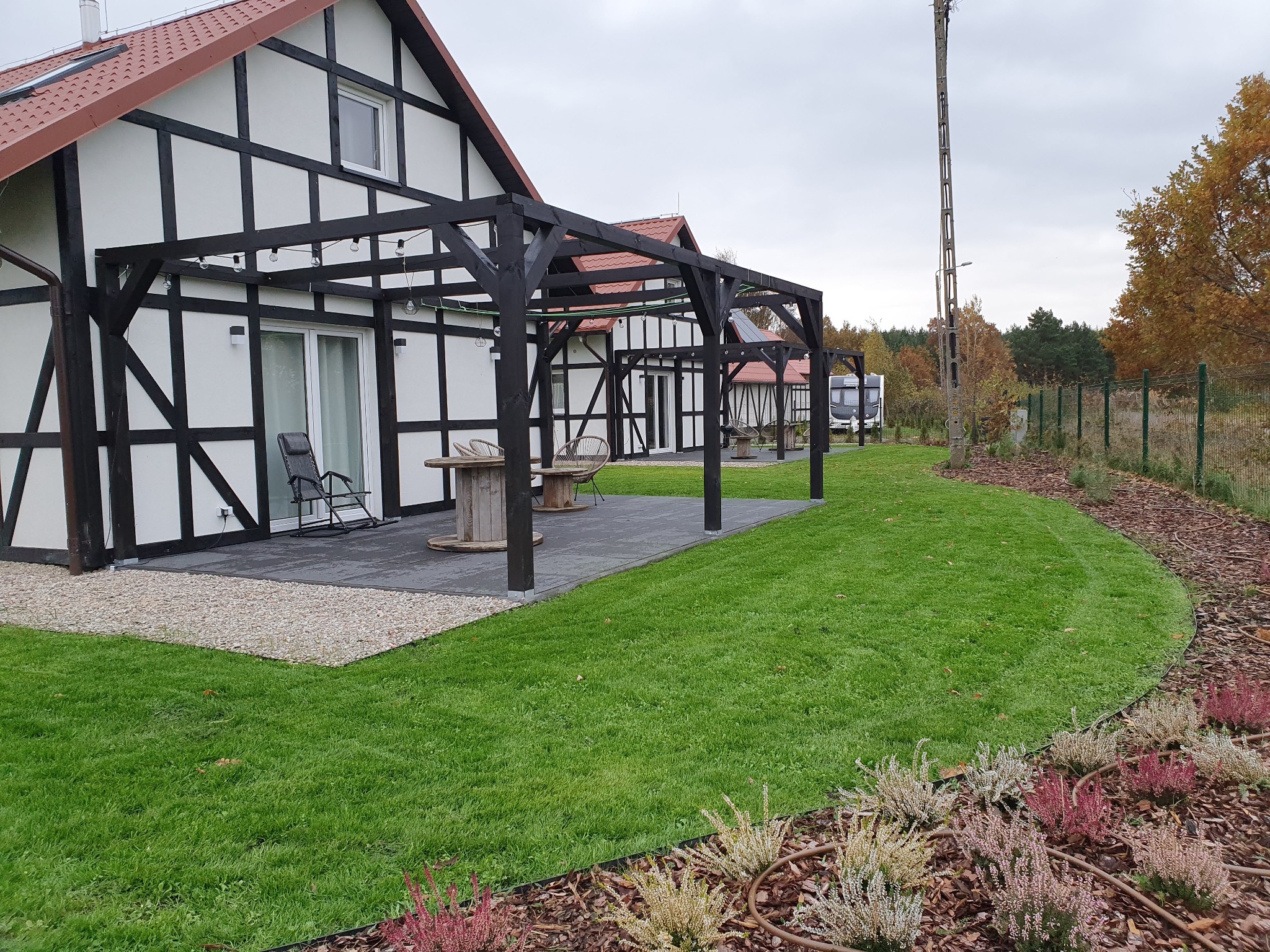 Ogród przydomowy z równo przystrzyżonym trawnikiem, rabatami z wrzosami i drewnianą pergolą z oświetleniem, na tle domu w stylu szachulcowym.