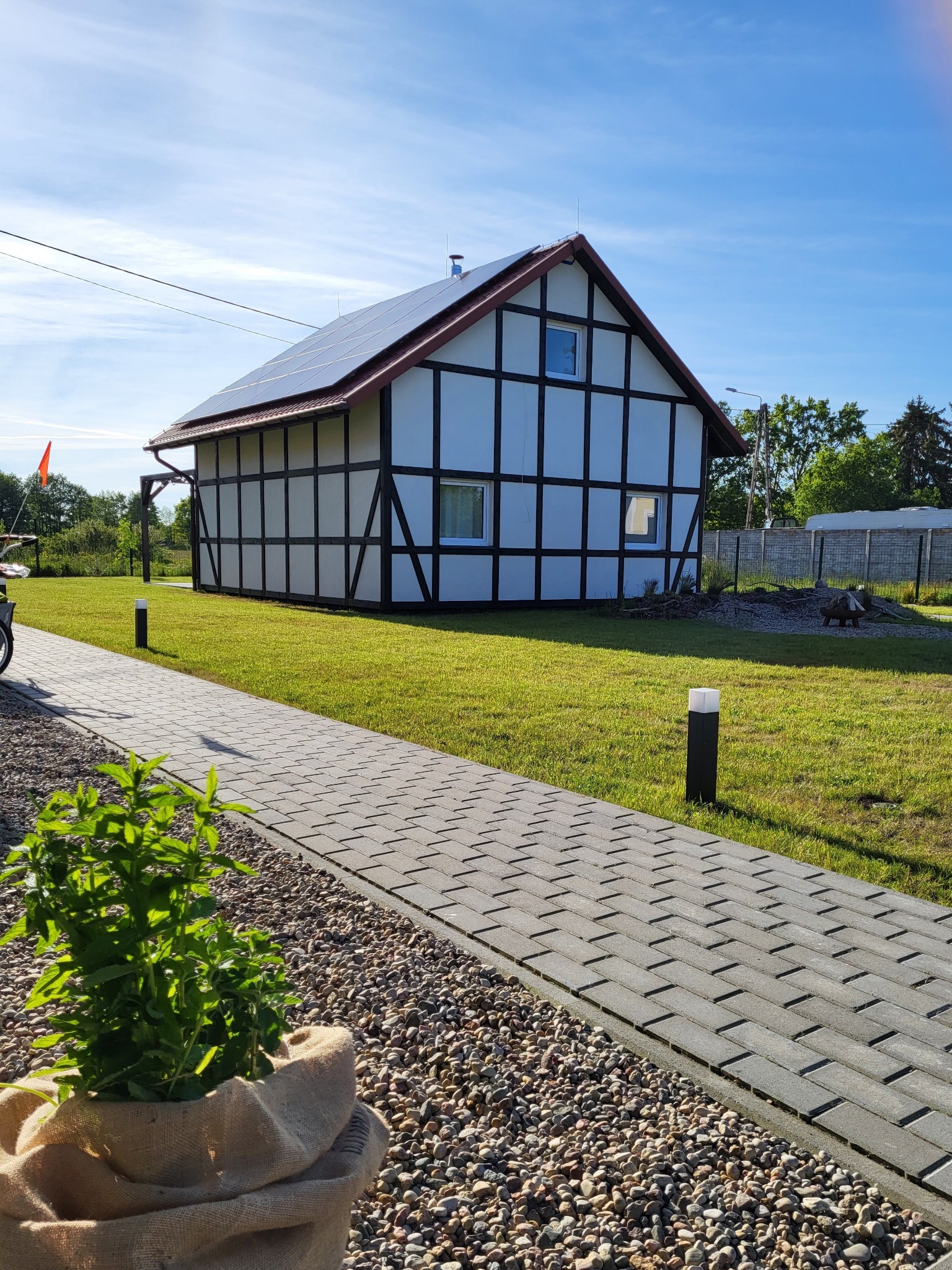 Nowoczesny dom z muru pruskiego z panelami słonecznymi na dachu, zadbany trawnik, ścieżka z kostki brukowej i roślina w jutowym worku na pierwszym planie.